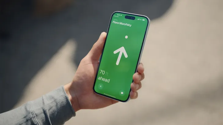 A hand holds and iPhone 15 which displays the Precision Finding feature. The screen is green with a white arrow pointing straight ahead.