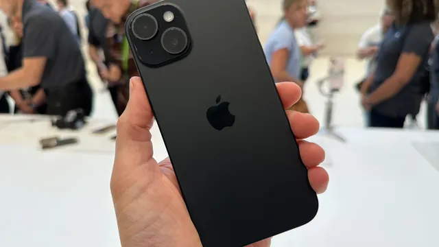 A hand holds a black iPhone 15, the back of the phone is facing the viewer. A blurred crowd is visible in the background.