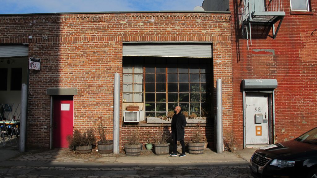 A person stands outside a red brick warehouse building in Red Hook, looking up at the architecture, with large factory windows, planters, and weathered industrial details. 