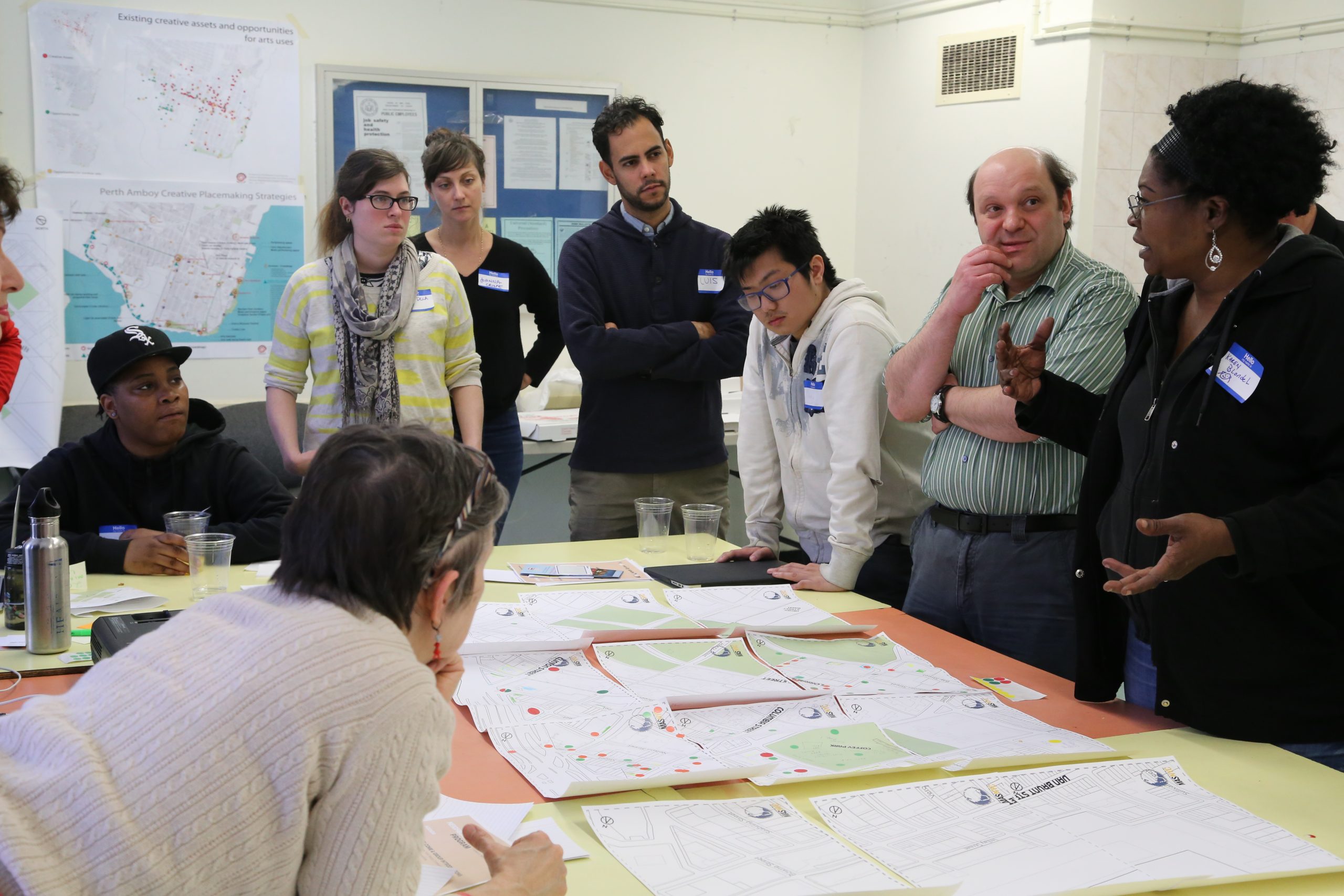 Group of people standing around a table covered with neighborhood maps marked with colored dots, gathered for a community planning workshop. Maps and creative placemaking strategy posters line the wall behind them. (
