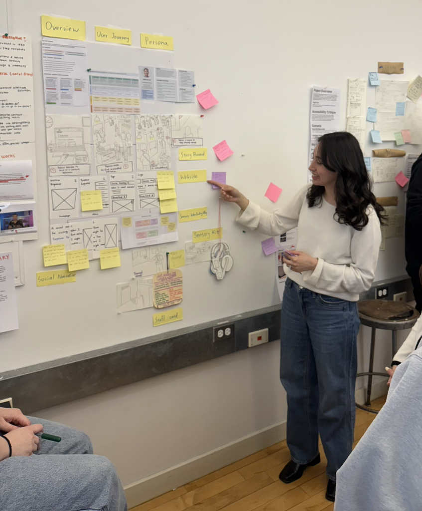 Alaa Shihab presenting the Design Progress Gallery: physical board displaying research synthesis, user journey map, persona, design sketches, and all four kit components. Photographed during class critique, April 2026. 
